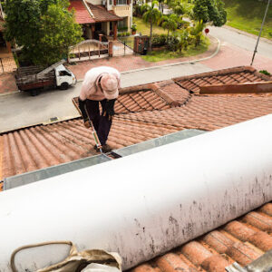 A worker on a rooftop applying sealant near a solar panel while inspecting potential signs of roof leaks, with a residential neighborhood visible in the background.